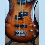 Detail těla elektrické basové kytary s lakovaným povrchem z dřeva s výraznou kresbou zrn. Nástroj má dva snímače typu humbucker v černé barvě a tři c…