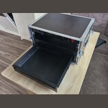 Černý transportní flightcase s hliníkovými hranami a vrškem. V horní části je osazen přístrojem s nápisy „L-Acoustics“ a „LA1I“. V dolní části je vys…