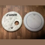 Dvě bílé bubnové kožehy položené na dřevěné podlaze. Levý kožeh s otvorem má nápis SONOR a menší štítek s textem SONOR, Natural Power. Pravý kožeh je…