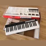 Bílá klávesová klaviatura s černým ovládacím panelem, na kterém je nápis iRig Keys I/O. Nástroj leží v otevřené krabici s polystyrenovými vložkami. V…