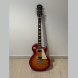 Elektrická kytara s trupem v červeně-žlutém sunburst provedení a bílým pickguardem. Na hlavě hmatníku je bílý nápis Epiphone a pod ním kurzivou Les P…