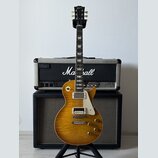 Elektrická kytara typu Les Paul s žlutým plamenovitým topem a bílým pickguardem. Na hlavě hmatníku je zlatý nápis Gibson. Kytara stojí na stojanu pře…