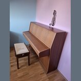Dřevěný pianino v hnědé barvě s doprovodnou stolečkou s béžovým čalouněním. Nástroj je umístěn v rohu místnosti u růžové a modré stěny. Na povrchu ná…