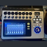 Digitální mixážní pult stříbrné barvy s černým rámem. V pravém horním rohu je nápis QSC TouchMix-8. Zařízení obsahuje osm vstupních kanálů s konektor…