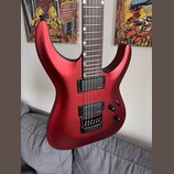 Elektrická kytara s červeným matným povrchem a hnědým hmatníkem. Nástroj má dva černé humbuckery a černý most s integrovaným pákovým systémem. Na těl…