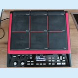 Černý sampler s devíti gumovými pady a červeným rámem. Na spodním ovládacím panelu je bílý nápis Roland SP-90 SX. Zařízení obsahuje displej s textem…