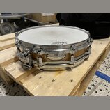 Na fotce je malý snare drum s dřevěným korpusem v přirozené hnědé barvě a chromovaným kováním. Na korpusu je viditelný oválný štítek s nápisy „Pearl“…