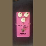 Růžový efekt pedál Analog Delay s třemi bílými oválnými knoflíky označenými jako DELAY TIME, REPEAT a BLEND. Na spodní části je nápis ANALOG DELAY, l…