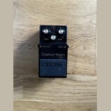 Černý efekt pedál s nápisy Distortion a DS-1. Na horní části jsou tři stříbrné ovládací knoflíky s popisky LEVEL, TONE a DIST, červená kontrolka CHEC…