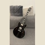 Černá polodublová elektrická kytara s bílým lemováním a černým pickguardem. Na hlavě hmatníku je viditelné logo Gibson. Nástroj má dva humbuckery, čt…