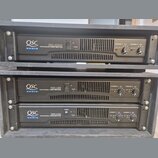 Tři černé rackové výkonové zesilovače značky QSC AUDIO. Horní jednotka nese nápis RMX 1850HD PROFESSIONAL POWER AMPLIFIER, prostřední RMX 1450 PROFES…