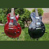 Dvě fotografie elektrických kytar typu Les Paul, jedna v červené a druhá v černé barvě. Obě mají dva humbuckery, tmavý hmatník a černý plechtrpickgua…