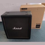 Černý kytarový reproduktor s logem Marshall uprostřed čelní síťovky. Box má hranatý tvar, černé potažení a zlatý lem kolem sítě. Vedle nástroje stojí…