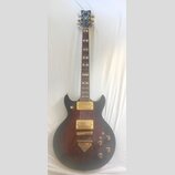 Elektrická kytara s tmavým, přechodovým povrchem od černé po hnědou a zlatými komponenty. Na hlavě je bílý nápis Ibanez a logo s ornamentem, hриф má…