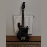 Černá elektrická kytara s černým pickguardem a hnedým hmatníkem. Na hlavě je bílý nápis "SAGA". Nástroj má dva snímače typu humbucker, tři ovládací k…