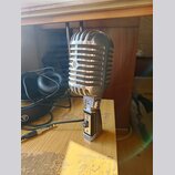 Stříbrný mikrofon s kovovou mřížkou v retro stylu, umístěný na stolním stojanu. Na přední straně mřížky je viditelné kulaté logo s písmenem „S“. Na s…