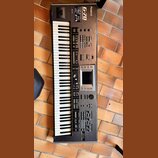 Tmavě šedý digitální klavír s bílými a černými klávesami. Na horní části je uveden nápis Roland a model G-70 MUSIC WORKSTATION. Přístroj obsahuje dis…