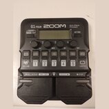 Černý multi-efektový procesor s nápisy G1 FOUR, ZOOM a Multi-Effects Processor. Zařízení obsahuje displej, čtyři otočné ovladače s popisky Lo, Mid, H…