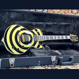 Elektrická kytara s jednovýřezem v tvrdém kufru. Tělo má výrazný žlutý a černý kruhový vzor, zlatý hardware a dva humbuckery. Na hlavě hmatníku je vi…