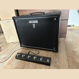 Černý kytarový kombozesilovač s logem BOSS a pod ním symbolem stylizovaného meče. Před zesilovačem je umístěn černý nožní spínač s nápisy BOOSTER, MI…