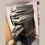 Stoh šedých akustických pěnových panelů s pyramidovou strukturou povrchu. Panely jsou naskládány na černém nábytkovém modulu a několika kusech je opř…