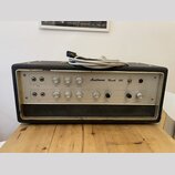 Černý kytarový kombozesilovač s bílým předním panelem a kovovým rámem. Na panelu je nápis Austrovox Reverb 100 a řada ovládacích knoflíků s popisky i…