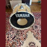 Světle béžový basový buben s černým předním koženem. Na koženě je bílý nápis YAMAHA a uprostřed je vyříznutý kruhový otvor. Nástroj má chromované lug…