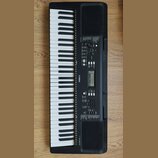 Černý digitální klavír s bílými a černými klávesami. Na korpusu je vlevo nápis PSR-E323 a uprostřed logo YAMAHA. Nástroj obsahuje ovládací panel s di…