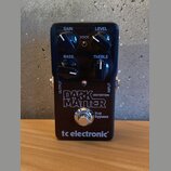 Černý efekt pedál s nápisy GAIN, LEVEL, BASS a TREBLE u ovládacích knoflíků a přepínačem VOICE. Uprostřed je velký bílý nápis DARK MATTER DISTORTION…