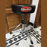 Černý polstrovaný bubenícký židun s chromovaným třínohým stativem. Na sedadle je bílo-červené logo s nápisem Gibraltar HARDWARE. Na černé části mecha…