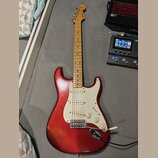 Červená elektrická kytara typu Stratocaster s bílým pickguardem a světle hnědým hmatníkem. Na hlavě nástroje je viditelné logo Fender. Kytara má tři…