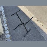 Černý kovový stojan na hudební nástroj s dvěma polstrovanými oporami. Stojan má sklapovací konstrukci a stojí na asfaltovém povrchu. Na fotce nejsou…