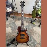 Elektrická kytara s trupem v hnědém sunburst provedení a viditnou strukturou dřeva. Na hlavě je bílé logo LTD a nápis Deluxe, na hmatníku jsou bílé v…