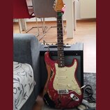 Elektrická kytara typu Stratocaster v červené barvě s výrazně odřeným povrchem a viditelným dřevem. Má krémový pickguard, tři snímače a chromovaný mo…
