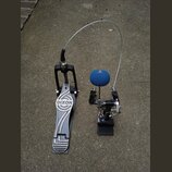 Černý basový pedál s kovovým šlapadlem, na kterém je bílý nápis DIXON. K pedálu vede flexibilní kovové táhlo k mechanismu u bubnu. Mechanismus je vyb…