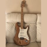 Elektrická kytara s přírodním dřevěným povrchem těla a hmatníkem. Na hlavě je viditelné logo Fender Stratocaster. Bílý pickguard obsahuje tři snímače…
