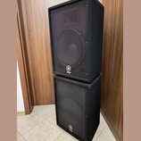 Dvě černé reproduktorové bedny v kostce, které jsou naskládány na sebe. Obě mají přední kovovou mřížku a v dolní části loga s nápisem YAMAHA. Povrch…