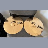 Dvě mosazné činely položené na šedém textilním povrchu. Horní činel má hladký povrch a nápis PAiste PSL 8 14" HI-HAT BOTTOM. Dolní činel má kovaný po…