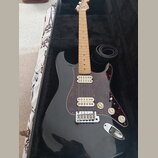 Černá elektrická kytara s tvarem typu Stratocaster, světlým hmatníkem a červeně vzorovaným pickguardem. Nástroj má tři snímače, tři ovládací knofíky…