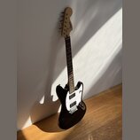 Černá elektrická kytara s bílým pickguardem a hnědým hmatníkem. Na hlavě nástroje je viditelné logo Squier. Kytara je opřena o bílou zeď na dřevěné p…
