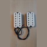 Dvě elektrické kytarové snímače typu humbucker s bílými plastovými kryty. Na obou je z levé strany svisle napsán nápis Seymour Duncan. Snímače mají č…