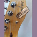 Detail hlavy elektrické kytary z přírodního dřeva. Na dřevě je viditelné částečné logo Fender. Na strunách je upevněn kovový stahovací kousek s nápis…