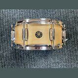 Fotografie znázorňuje snare drum s dřevěným korpusem v přírodní světle barvě a chromovaným obručemi. Uprostřed korpusu je umístěno kulaté logo s nápi…