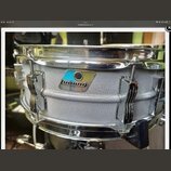 Stříbrný snare drum s texturovaným povrchem a chromovanými obručemi. Na korpusu je viditelný štítek s logem ludwig a nečitelným nápisem pod ním. Nást…