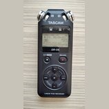 Černý digitální záznamový systém s dvěma stříbrnými mikrofony v horní části označenými písmeny L a R. Na přístroji je bílý nápis TASCAM, modelové ozn…