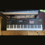 Černý digitální klavír s bílými a černými klávesami. Na pravé straně je nápis Roland JUNO-DS a modrá sekce s označením PHRASE PAD. V horní části jsou…