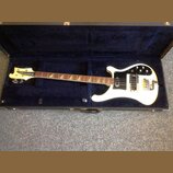 Bílá elektrická basová kytara v černém tvrdém kufru s modrým polstrováním. Na hlavě nástroje je nápis Rickenbacker. Kytara má bílý pickguard, dva sní…