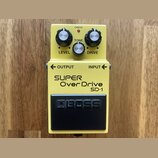 Žlutý efekt pedál s černým spínacím tlačítkem. Na spodní části je nápis SUPER Over Drive SD-1 a na spínači logo BOSS. V horní části jsou tři ovládací…