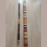 Dvě didgeridou z dřeva s různým povrchovým zdobením. Levý nástroj je tmavě hnědý s barevnými ornamenty a vzory. Pravý nástroj je světlejší, zdobený h…