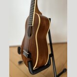 Hnědý akustický nástroj typu ukulele s dřevěným povrchem a světlým lemováním. Nástroj je umístěn v černém stojanu. Na viditelné části nejsou žádné zn…